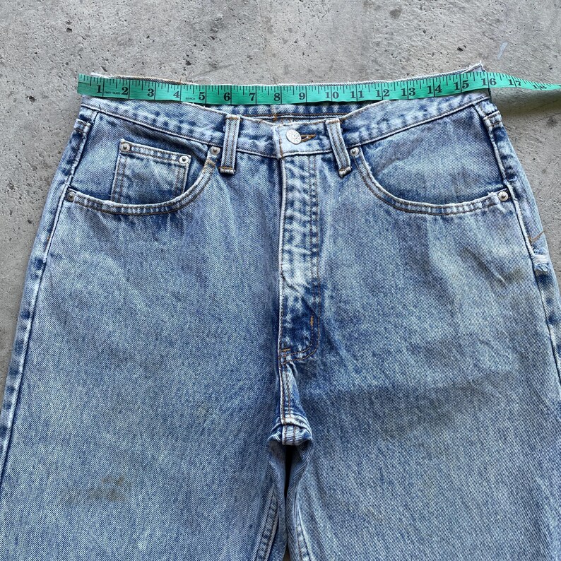 May include: Light blue denim jeans with a classic five-pocket design. The jeans have a button and zipper closure, belt loops, and a faded wash. A measuring tape is placed across the waist, showing a measurement of approximately 15 inches.