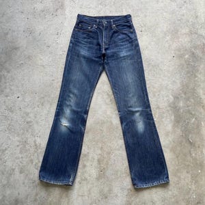 May include: Dark wash denim jeans with a classic five-pocket design. The jeans show signs of wear and tear, including a distressed area on the left knee. The jeans have a straight leg fit.