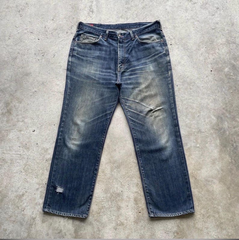K&ouml;nnte beinhalten: Verblasste blaue Jeans mit klassischem F&uuml;nf-Taschen-Design. Die Jeans weisen Gebrauchsspuren auf, einschlie&szlig;lich Abnutzung und einem kleinen Riss am linken Bein. Die Jeans haben einen geraden Bein-Schnitt und einen Knopf- und Rei&szlig;verschluss.