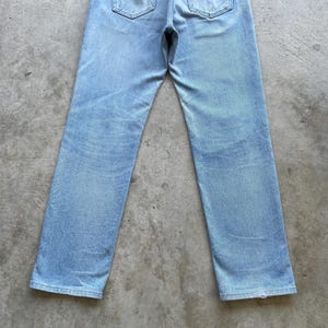 May include: Light blue denim jeans with a classic five-pocket design. The jeans have a straight leg cut and a faded wash, giving them a vintage look. The back pockets and waistband are visible.