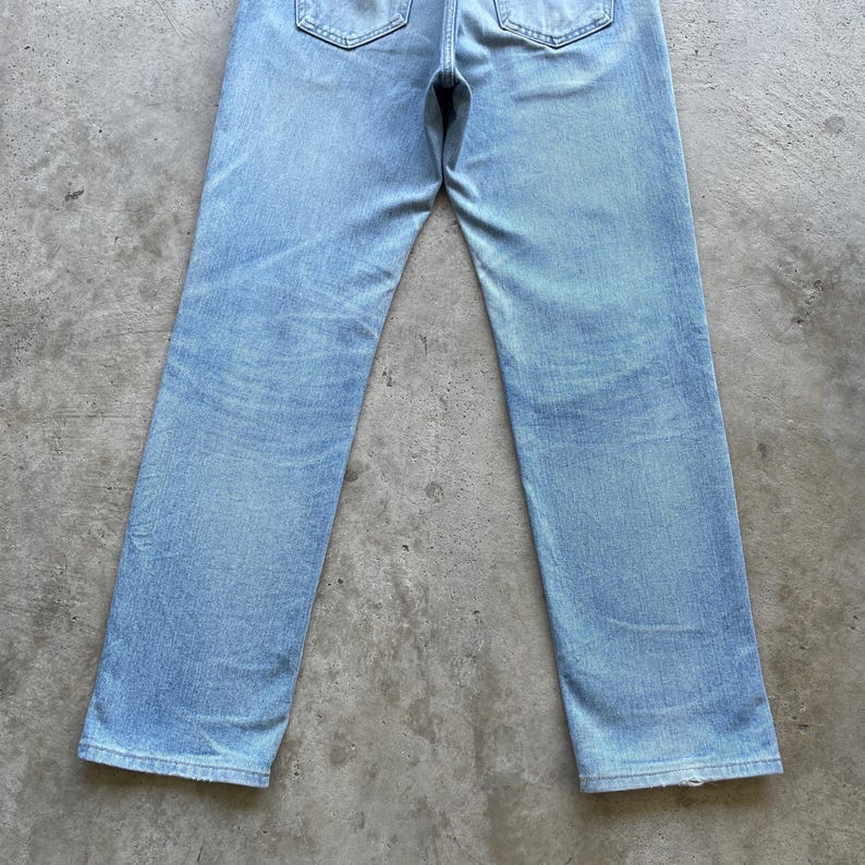 May include: Light blue denim jeans with a classic five-pocket design. The jeans have a straight leg cut and a faded wash, giving them a vintage look. The back pockets and waistband are visible.