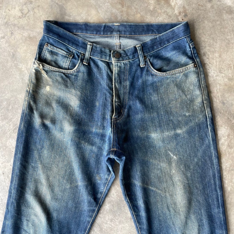 Puede incluir: Vaqueros de mezclilla azul descolorido con cierre de bot&oacute;n y dise&ntilde;o cl&aacute;sico de cinco bolsillos. Los vaqueros muestran signos de desgaste, con &aacute;reas desgastadas y parches de color claro, d&aacute;ndoles un aspecto vintage. Los vaqueros son de lavado medio.