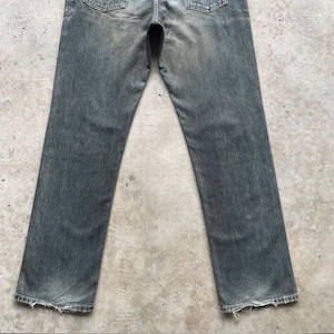 May include: Faded blue denim jeans with a straight leg cut. The jeans show signs of wear and tear, with distressed edges at the bottom of the legs. The back pockets and waistband are visible.