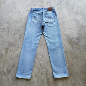 May include: Light blue denim jeans with a classic five-pocket design. The jeans have a faded wash and a straight leg cut. A brown leather patch is visible at the back waistband. The jeans are laid flat on a concrete surface.