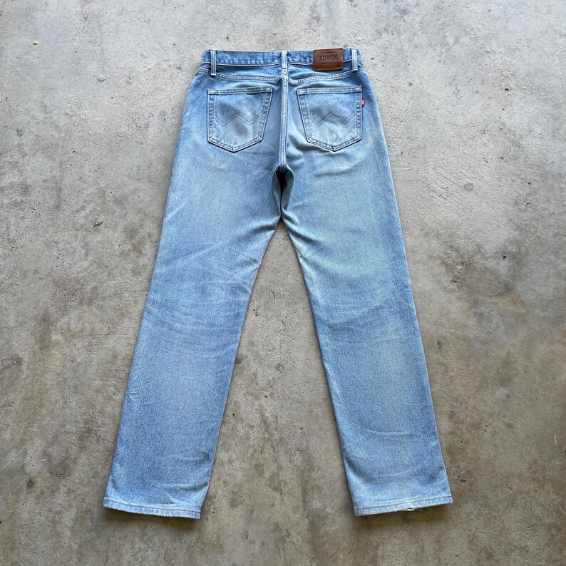 May include: Light blue denim jeans with a classic five-pocket design. The jeans have a faded wash and a straight leg cut. A brown leather patch is visible at the back waistband. The jeans are laid flat on a concrete surface.
