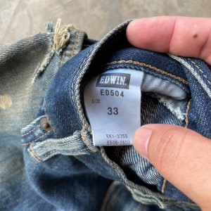Puede incluir: Primer plano de unos vaqueros de mezclilla azules con una etiqueta EDWIN. La etiqueta muestra "ED504" y la talla "33". Los vaqueros muestran signos de desgaste y detalles desgastados.