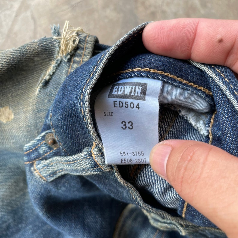 Puede incluir: Primer plano de unos vaqueros de mezclilla azules con una etiqueta EDWIN. La etiqueta muestra "ED504" y la talla "33". Los vaqueros muestran signos de desgaste y detalles desgastados.