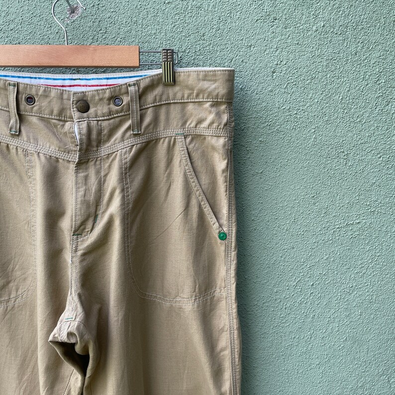 May include: Khaki-coloured trousers with a button and zip fastening, featuring front and back pockets. The trousers have a straight leg design and are hanging on a wooden hanger. The waistband has a striped detail with red, white, and blue colours.