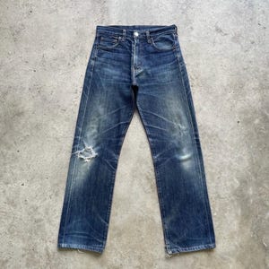 May include: Vintage blue denim jeans with a distressed look. The jeans have a button fly, classic five-pocket design, and a ripped knee. The denim has faded areas and wear marks, giving them a worn-in appearance. The jeans are laid flat on a concrete surface.