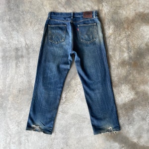 Puede incluir: Vaqueros azules descoloridos con un aspecto desgastado. Los vaqueros tienen un dise&ntilde;o cl&aacute;sico de cinco bolsillos, con costuras curvas distintivas en los bolsillos traseros. Una peque&ntilde;a etiqueta roja es visible en el bolsillo trasero y un parche de cuero marr&oacute;n en la cintura.