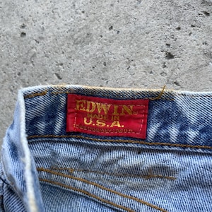 May include: Close-up of a section of blue denim jeans. A red rectangular tag is sewn onto the jeans, displaying the brand name "EDWIN" in gold lettering, with "MADE IN U.S.A." below it. The jeans have visible stitching.