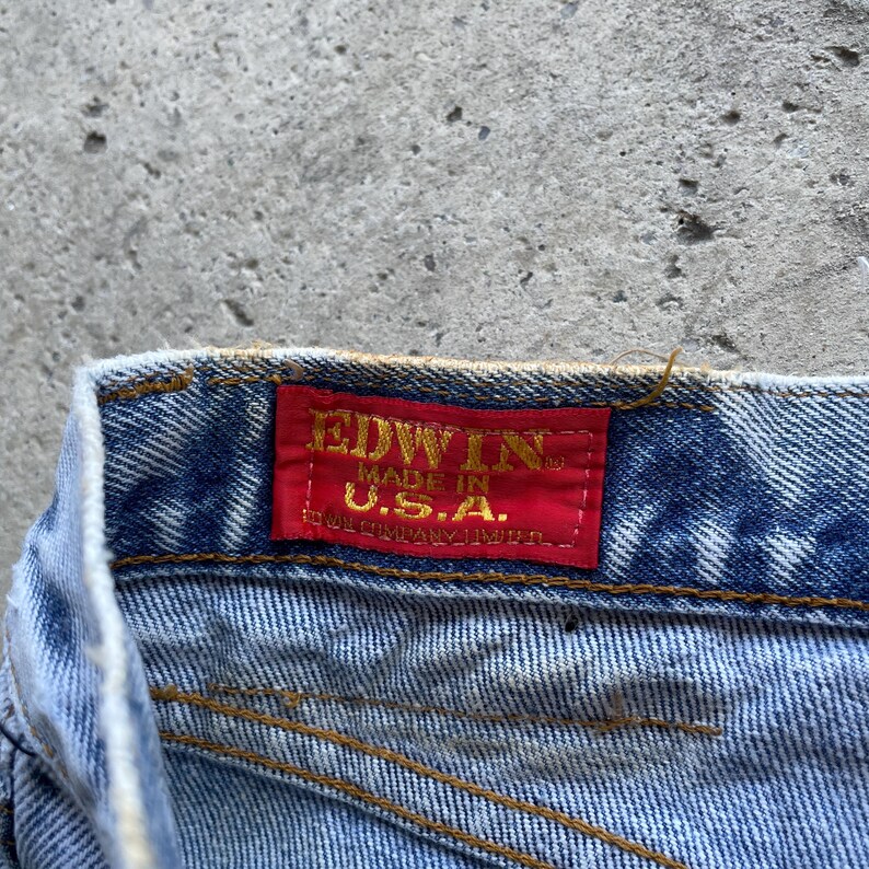 May include: Close-up of a section of blue denim jeans. A red rectangular tag is sewn onto the jeans, displaying the brand name "EDWIN" in gold lettering, with "MADE IN U.S.A." below it. The jeans have visible stitching.