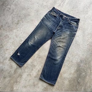 K&ouml;nnte beinhalten: Verblasste blaue Jeans mit einem Used-Look. Die Jeans haben ein klassisches F&uuml;nf-Taschen-Design, einen Knopfverschluss und ein abgenutztes Aussehen mit sichtbaren Ausbleichungen und einem kleinen Riss in der N&auml;he des Saums. Die Jeans liegen flach auf einer Betonoberfl&auml;che.