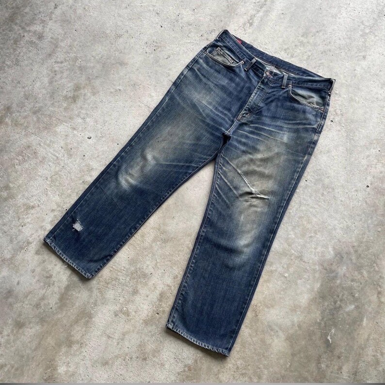 K&ouml;nnte beinhalten: Verblasste blaue Jeans mit einem Used-Look. Die Jeans haben ein klassisches F&uuml;nf-Taschen-Design, einen Knopfverschluss und ein abgenutztes Aussehen mit sichtbaren Ausbleichungen und einem kleinen Riss in der N&auml;he des Saums. Die Jeans liegen flach auf einer Betonoberfl&auml;che.