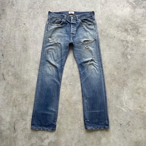 May include: Faded blue denim jeans with distressed details, including rips and wear marks. The jeans have a button fly and classic five-pocket design. The jeans are laid flat on a concrete surface.