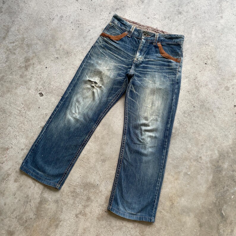 May include: Faded blue denim jeans with distressed details, including a tear on the left leg. Features brown leather accents on the front pockets and a worn waistband. The jeans have a classic five-pocket design.