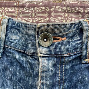 May include: Close-up of blue denim jeans with a button closure. The button is a tarnished silver color with the word "Edwin" around the edge. The jeans have a brown patterned lining and orange stitching.