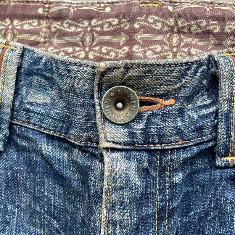 May include: Close-up of blue denim jeans with a button closure. The button is a tarnished silver color with the word "Edwin" around the edge. The jeans have a brown patterned lining and orange stitching.