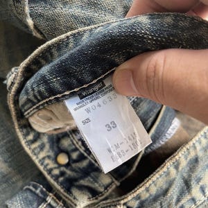 May include: Close-up of a pair of blue denim jeans with a faded wash. A white tag displays the Wrangler logo, size 33, and other details. The jeans show wear and tear.