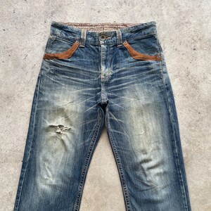 May include: Faded blue denim jeans with distressed details, including a ripped knee. Features brown suede-like pocket accents and a patterned waistband. The jeans have a button closure and classic five-pocket design.