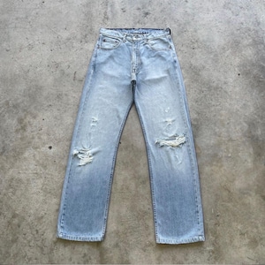 May include: Light blue denim jeans with distressed details, including rips at the knees. The jeans have a classic five-pocket design and a straight leg fit. The jeans are laid flat on a concrete surface.