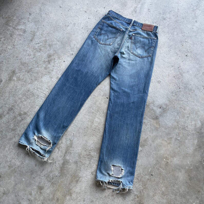 Pu&ograve; includere: Jeans di denim blu sbiadito con dettagli usurati, tra cui ginocchia strappate e orli sfilacciati. I jeans presentano un classico stile a cinque tasche, una toppa in pelle marrone e una chiusura con bottone e cerniera. I jeans sono appoggiati su una superficie di cemento.