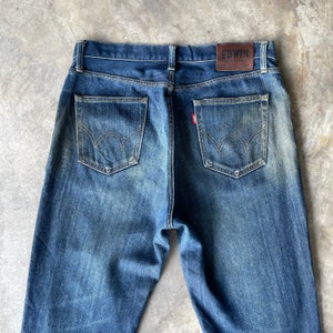 Puede incluir: Vaqueros de mezclilla azul con un dise&ntilde;o cl&aacute;sico de cinco bolsillos. Los bolsillos traseros presentan una costura curva distintiva. Un parche de cuero marr&oacute;n con la marca "EDWIN" es visible en la cintura. Una peque&ntilde;a etiqueta roja est&aacute; en el bolsillo trasero.