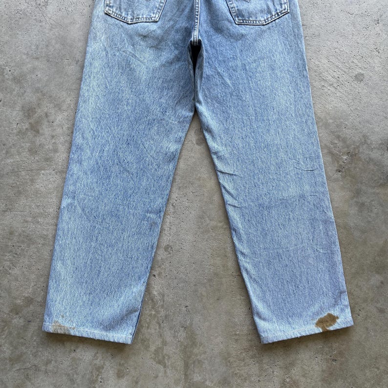 May include: Light blue denim jeans with a faded wash and a straight leg fit. The jeans have two back pockets and show signs of wear, including discoloration near the hem. The jeans are a classic style.