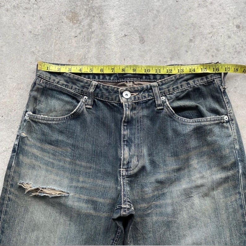 May include: Close-up of distressed blue denim jeans with a button closure and front pockets. A yellow measuring tape shows a waist measurement of approximately 18 inches. The jeans have a worn, faded appearance.