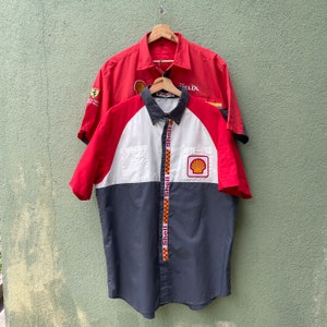 May include: Two short-sleeved button-down shirts hanging on wooden hangers. The front shirt is red, white, and navy blue with Shell branding. The back shirt is solid red with Ferrari and Shell logos. Both shirts are made of a durable fabric.