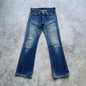 May include: Faded blue denim jeans with distressed details, including knee rips. The jeans have a classic five-pocket design, a button and zipper closure, and a slight flare at the hem.