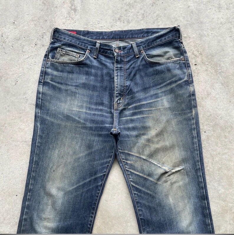 K&ouml;nnte beinhalten: Verblasste blaue Jeans mit einem abgenutzten Aussehen. Die Jeans haben ein klassisches F&uuml;nf-Taschen-Design, einen Knopfverschluss und sichtbare Abnutzungserscheinungen, einschlie&szlig;lich eines Risses am rechten Bein. Die Jeans haben einen Vintage-Look.