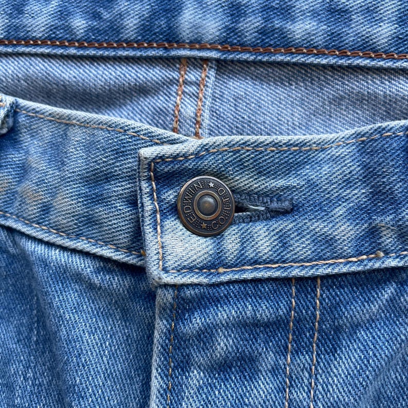 May include: Close-up of a pair of blue denim jeans. The image highlights the button closure, featuring a metal button with the text "NIMDEO CO." The jeans show signs of wear and tear, with visible stitching and a faded appearance.