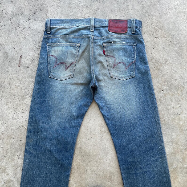 Puede incluir: Vaqueros de mezclilla azul descolorido con costuras rojas en los bolsillos traseros y un parche de cuero rojo. Los vaqueros tienen un dise&ntilde;o cl&aacute;sico de cinco bolsillos y una apariencia desgastada, lo que sugiere un estilo vintage o desgastado.