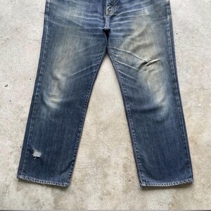 K&ouml;nnte beinhalten: Verblasste blaue Jeans mit Used-Details. Die Jeans hat einen geraden Beinverlauf, einen Knopfverschluss und sichtbare Abnutzungserscheinungen, darunter ein Riss am rechten Knie und ein Loch am linken Bein. Der Denim hat einen abgenutzten, Vintage-Look.