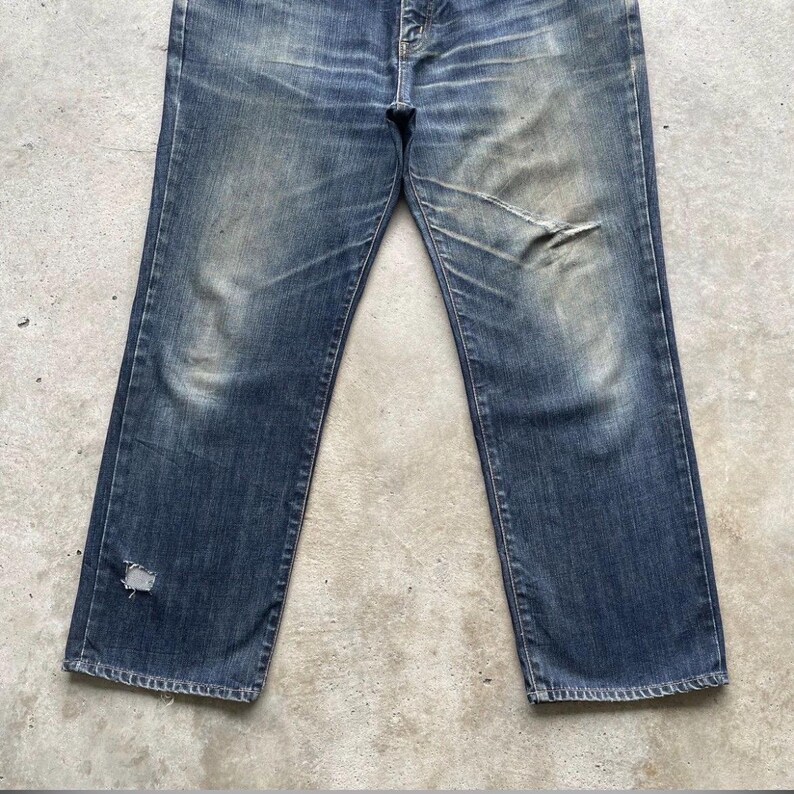 K&ouml;nnte beinhalten: Verblasste blaue Jeans mit Used-Details. Die Jeans hat einen geraden Beinverlauf, einen Knopfverschluss und sichtbare Abnutzungserscheinungen, darunter ein Riss am rechten Knie und ein Loch am linken Bein. Der Denim hat einen abgenutzten, Vintage-Look.