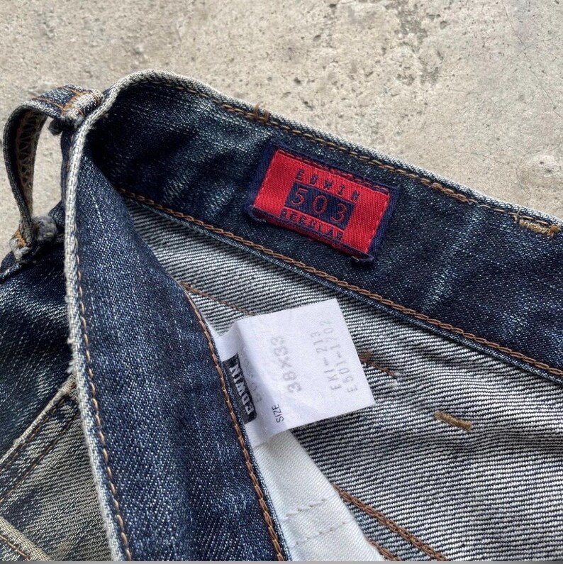 K&ouml;nnte beinhalten: Nahaufnahme einer dunkelblauen Edwin 503 Regular Jeans. Die Jeans hat ein rotes Etikett mit wei&szlig;em Text "Edwin 503 Regular". Ein wei&szlig;es Etikett zeigt die Gr&ouml;&szlig;e 38x33 und weitere Informationen. Die Jeans ist aus Denim.