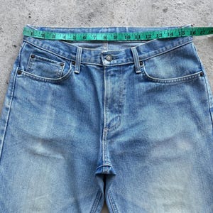 May include: Light blue denim jeans with a button closure and classic five-pocket design. A green measuring tape is laid across the waistband, showing a measurement of approximately 16 inches. The jeans have a faded wash and visible wear.