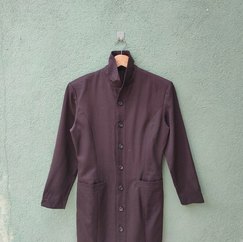 May include: A dark brown, long-sleeved coat with a high collar and a row of buttons down the front. The coat has two pockets and is hanging on a wooden hanger.