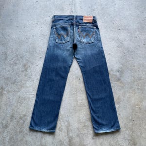 May include: Rear view of blue denim jeans with a classic five-pocket design. The jeans display the Levi's stitching on the back pockets, a red tab, and a brown leather patch. They have a straight leg cut and a faded wash, typical of vintage denim.