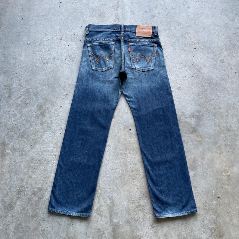 May include: Rear view of blue denim jeans with a classic five-pocket design. The jeans display the Levi's stitching on the back pockets, a red tab, and a brown leather patch. They have a straight leg cut and a faded wash, typical of vintage denim.