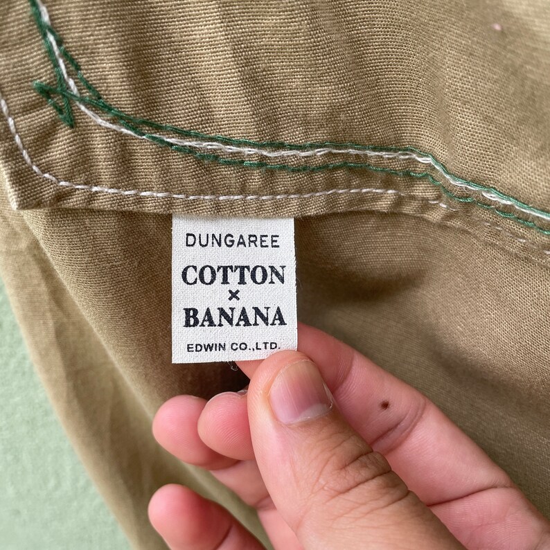 May include: Close-up of a tan fabric with a white label. The label reads "DUNGAREE COTTON x BANANA EDWIN CO., LTD." The fabric has white and green stitching along the edge. The fabric is held by a person's hand.