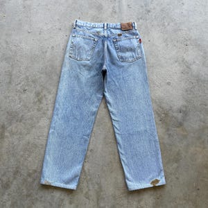 May include: Light blue denim jeans with a faded wash. The jeans have a classic five-pocket design, a brown leather patch, and a red tag. The back pockets feature a distinctive stitched design. The jeans are laid flat on a concrete surface.