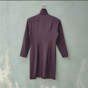 May include: A dark purple dress with long sleeves and a high collar, hanging on a metal hanger. The dress has a tailored fit and a simple, elegant design. The background is a light green wall.
