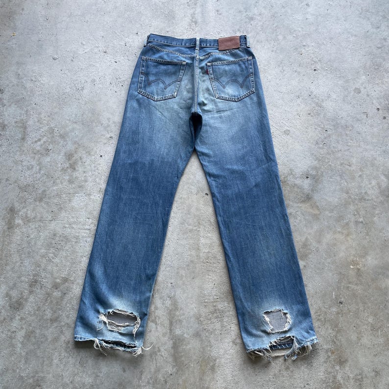 Pu&ograve; includere: Jeans di denim blu sbiadito con dettagli usurati, tra cui ginocchia strappate e un aspetto vissuto. I jeans presentano il classico stile a cinque tasche, una chiusura con bottone e cerniera e una toppa in pelle marrone sul retro.