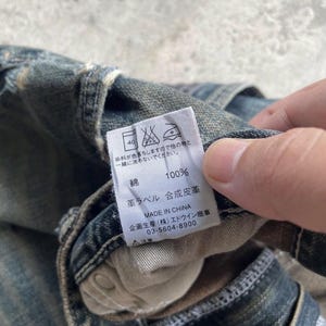 May include: Close-up of a denim garment with a white tag. The tag displays washing instructions, the word "cotton" and "100%", and other Japanese text. The garment is faded blue.