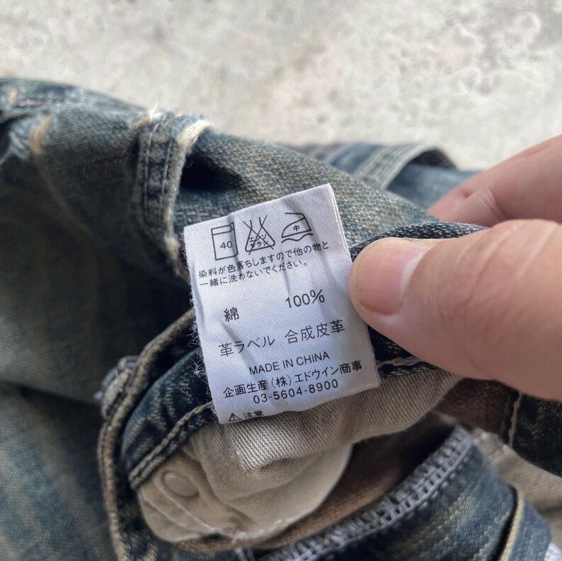 May include: Close-up of a denim garment with a white tag. The tag displays washing instructions, the word "cotton" and "100%", and other Japanese text. The garment is faded blue.