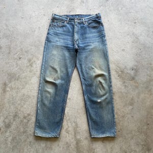 May include: Faded blue denim jeans with a button fly. The jeans show signs of wear and tear, with faded areas and a small hole near the knee. The jeans are a classic, straight-leg style.