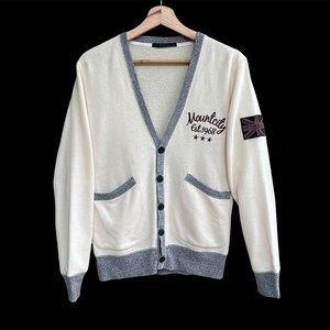 May include: Cream cardigan with grey trim, pockets, and buttons. It has the text "Mountcity Est.1968" and a Union Jack patch on the sleeve. The sweater is displayed on a wooden hanger against a black background.