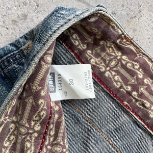 May include: Close-up of blue denim jeans with a patterned interior lining. The jeans have a size 30 label and the brand name "Edwin". The lining features a repeating arrow design.